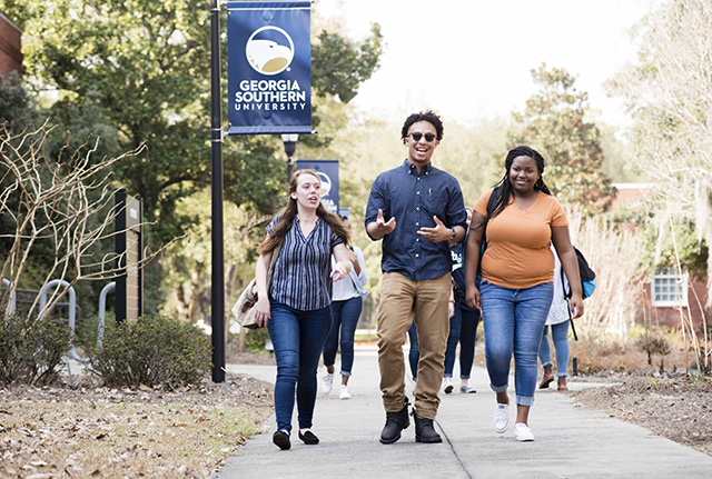 A group of students walk along a campus sidewalk near a “Georgia Southern University” banner, talking and smiling on a sunny day with trees and buildings in the background.