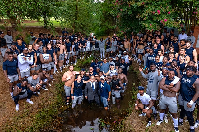 A large group of football players and coaches pose together outdoors, surrounding a small stream. Many are in athletic gear, some shirtless, and coaches in suits stand in the water.