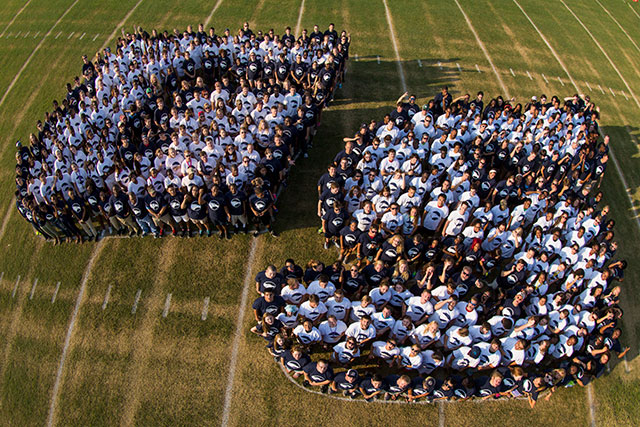 A large group of people wearing white and dark shirts stand on a grassy field, arranged to form GS when viewed from above.