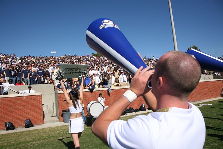 A cheerleader in white holds up a sign while another person shouts through a blue megaphone toward a crowded stadium of spectators on a sunny day.