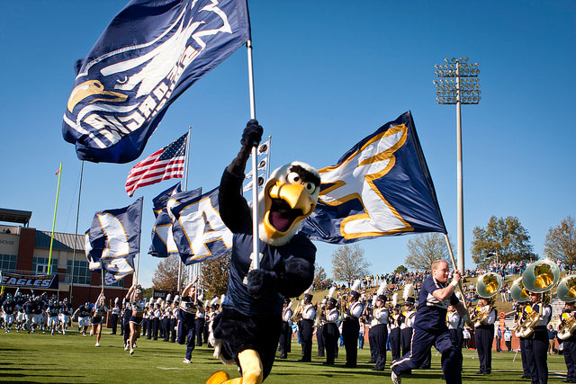 Gus waves a large flag on a football field, leading a marching band and cheerleaders. Other large flags and an American flag are in the background, with a crowd watching from the stands.