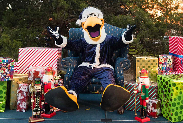 Gus sits in a festive chair, surrounded by wrapped presents and toy nutcrackers, with trees in the background. The mascot has its arms open and looks cheerful.