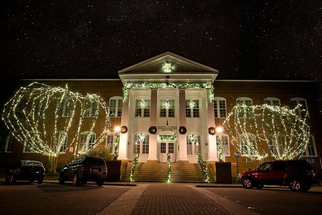 A large building with columns is decorated with white holiday lights on the trees and railings at night. Three wreaths hang on the doors, and several cars are parked in front under a starry sky.