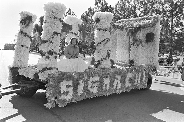 A woman in a formal dress sits on a parade float covered in flowers. The float is on a street lined with trees, and it appears to be part of a celebratory procession.