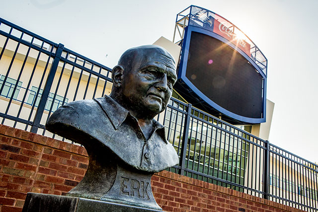 A bronze statue of Erk Russell sits in front of a large, unlit scoreboard and fence at an outdoor sports stadium, with sunlight shining from behind the scoreboard.