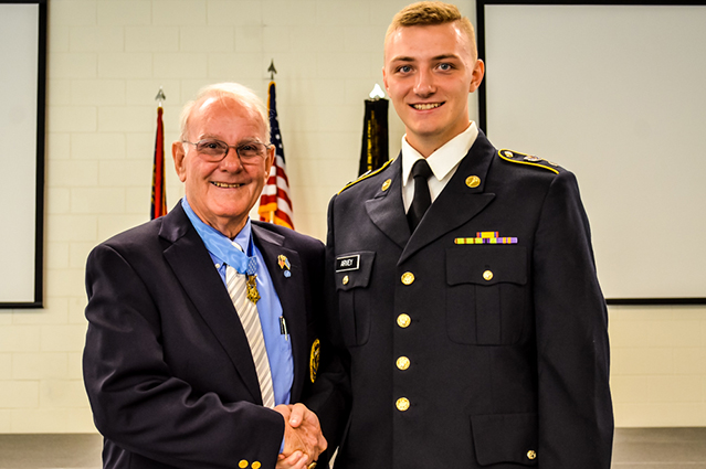 An older man in a suit shakes hands with a young man in a military uniform. Both are smiling. American flags are visible in the background.