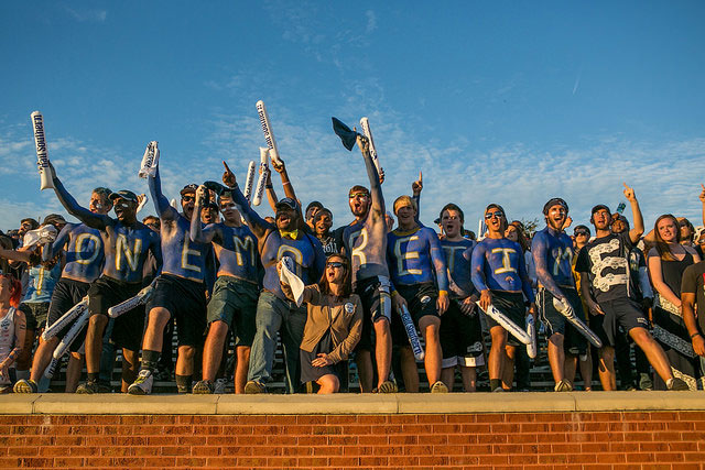 A group of enthusiastic sports fans, many with painted chests spelling “ONE MORE TIME,” cheer and wave foam sticks in a stadium under a clear blue sky.