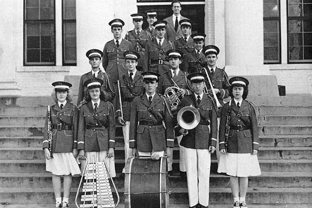A vintage black-and-white photo of a school marching band, with boys and girls in uniform holding instruments, posing on the steps of a building with large columns and windows behind them.