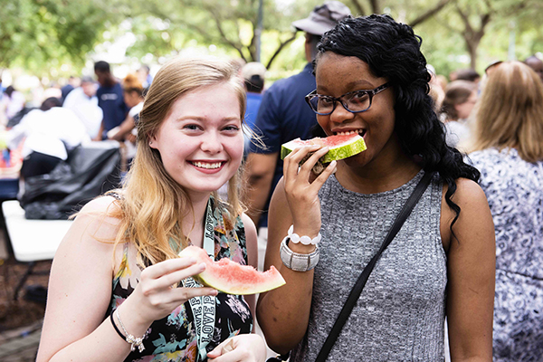 Two young women stand outdoors in a crowd, smiling and enjoying slices of watermelon. One wears a floral top and the other a gray dress.