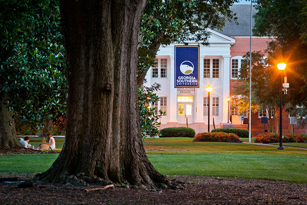 A large tree stands in the foreground on a grassy lawn, with students sitting nearby. In the background, a building with a Georgia Southern University banner is lit by street lamps at dusk.