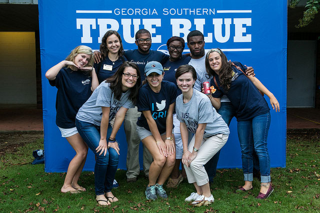 A group of nine smiling young adults pose together in front of a blue banner that reads Georgia Southern True Blue. They are outdoors and wearing casual clothing, most in navy blue shirts.