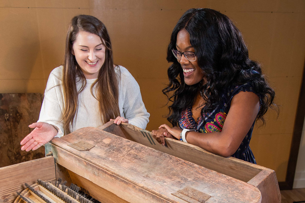Two women smile and examine the inside of a large, old wooden chest or box with compartment dividers; one gestures toward the contents while the other looks on with interest.