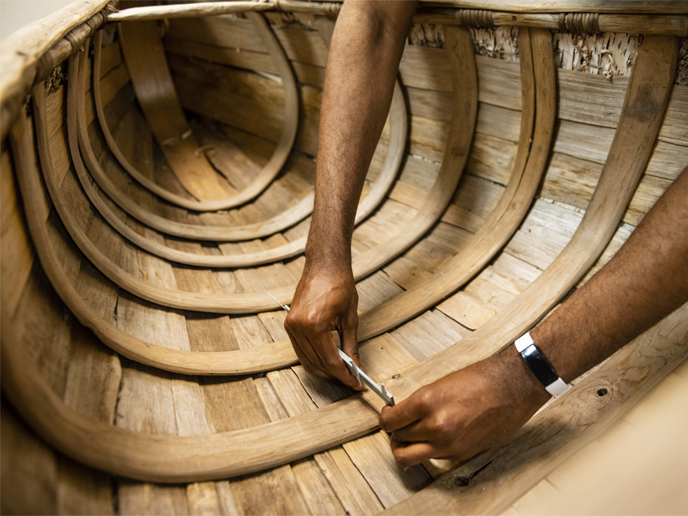 Close-up of a person’s hands working inside a wooden boat, using a tool to adjust or assemble the curved wooden ribs and planks of the boat’s interior structure.