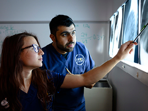 Two healthcare professionals, one woman and one man, examine and discuss X-ray images illuminated on a lightbox. The man points at the X-rays while the woman attentively observes, both wearing dark blue scrubs.