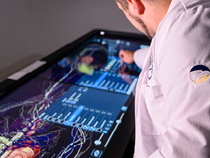 A person in a lab coat interacts with a large touchscreen displaying a detailed anatomical diagram and data, possibly using it for medical or educational purposes.