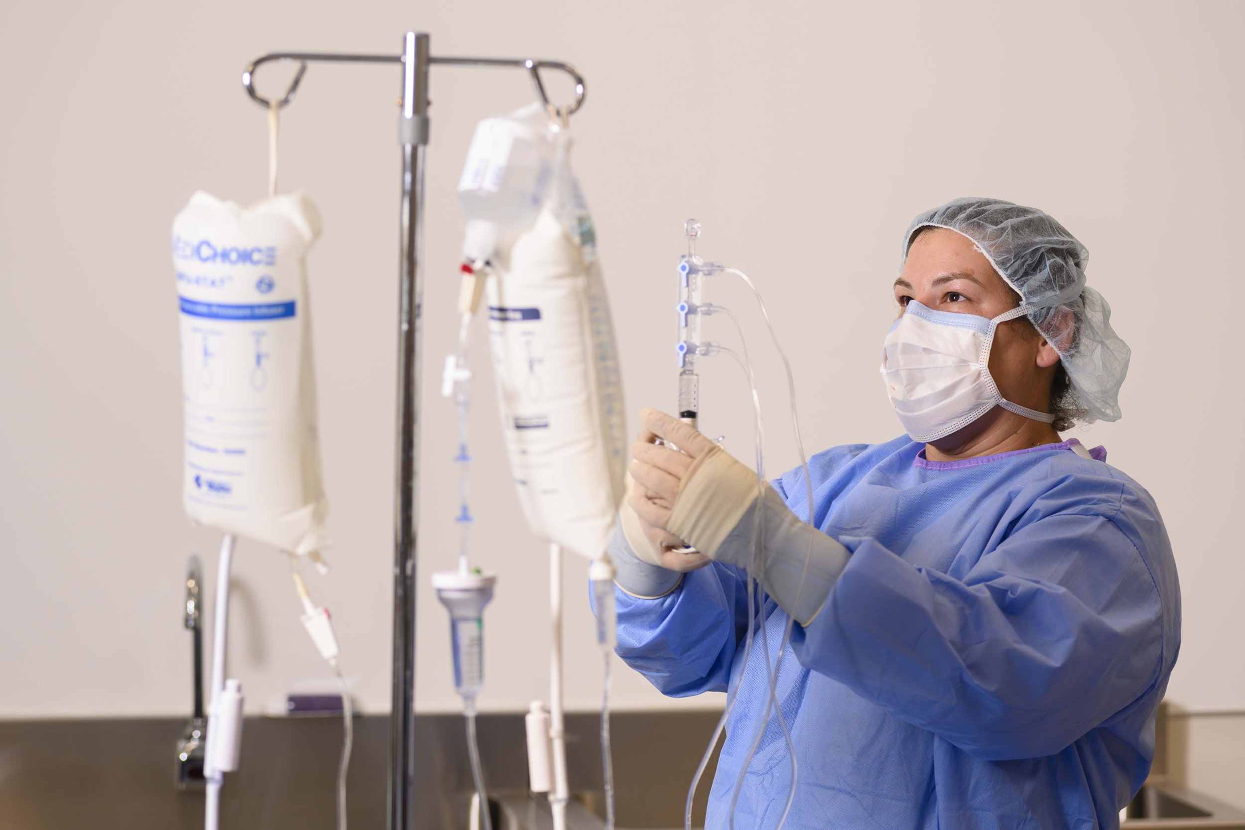 A healthcare worker wearing a mask, hair cap, gloves, and gown adjusts IV bags and tubing on a medical stand in a clinical setting.