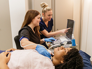 Two medical professionals perform an ultrasound on a patient’s neck. One operates the ultrasound probe while the other points at the monitor displaying the results. The patient lies on a bed, looking relaxed.
