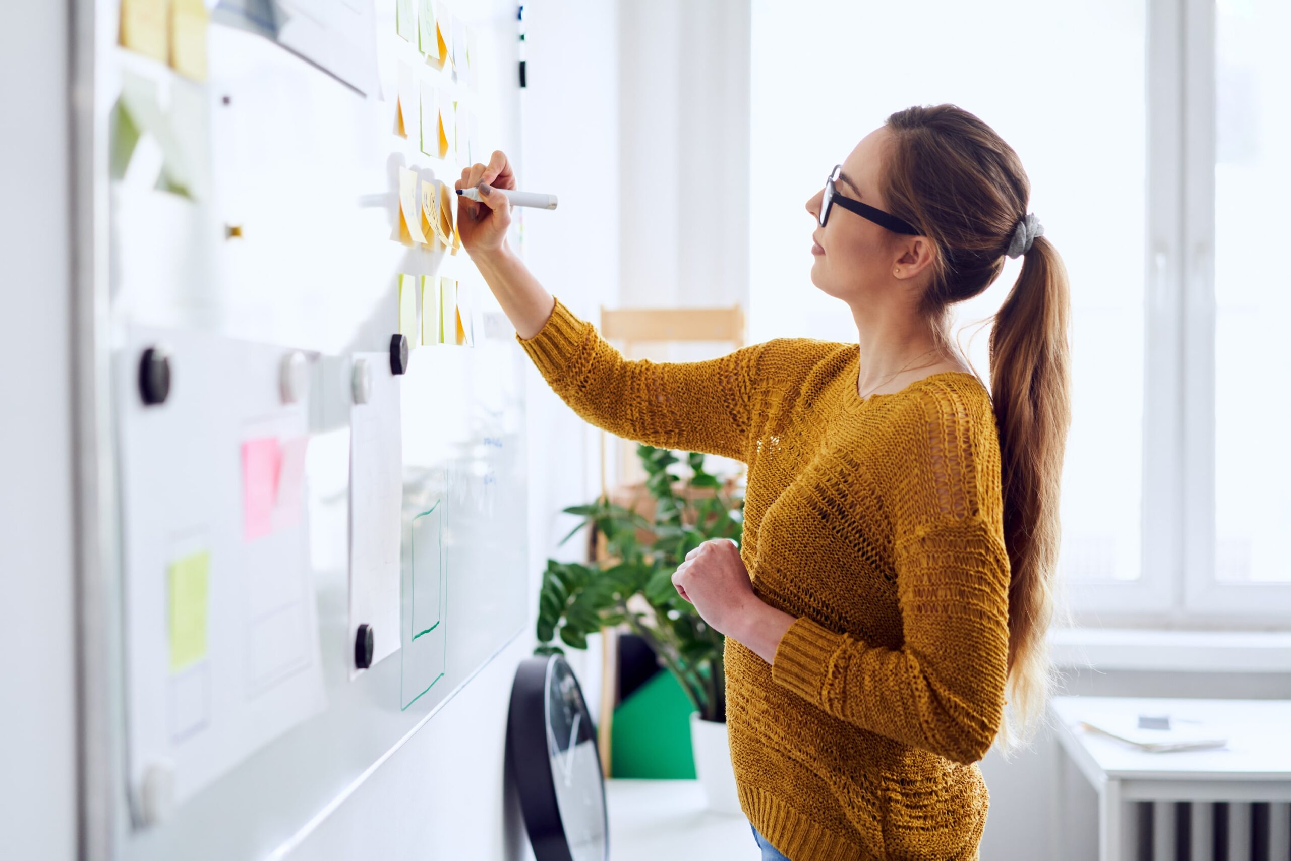 A woman with glasses and a ponytail, wearing a yellow sweater, writes on a whiteboard covered with sticky notes in a bright office space with a plant and large window in the background.