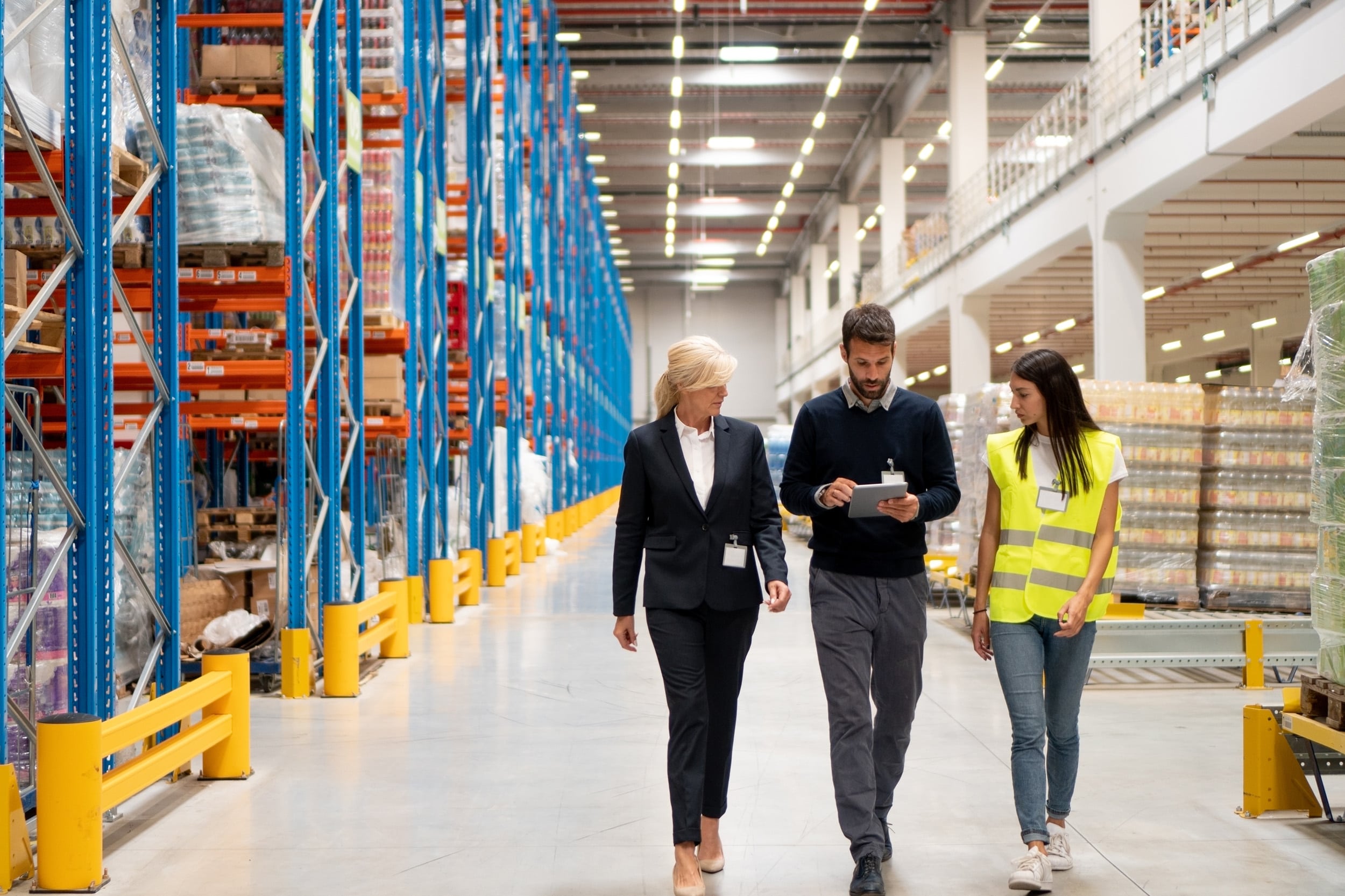 Three people walk through a warehouse with high shelves filled with boxes; one woman wears a suit, one man looks at a clipboard, and another woman wears a yellow safety vest and jeans.