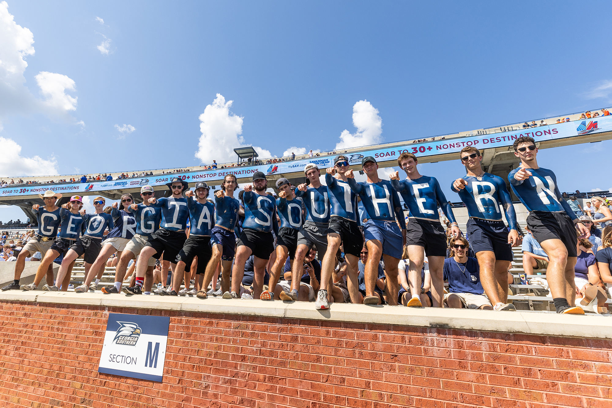 A group of people at a stadium stand in a row, each with a painted on large letter to spell GEORGIA SOUTHERN. Spectators fill the background stands under a partly cloudy sky.