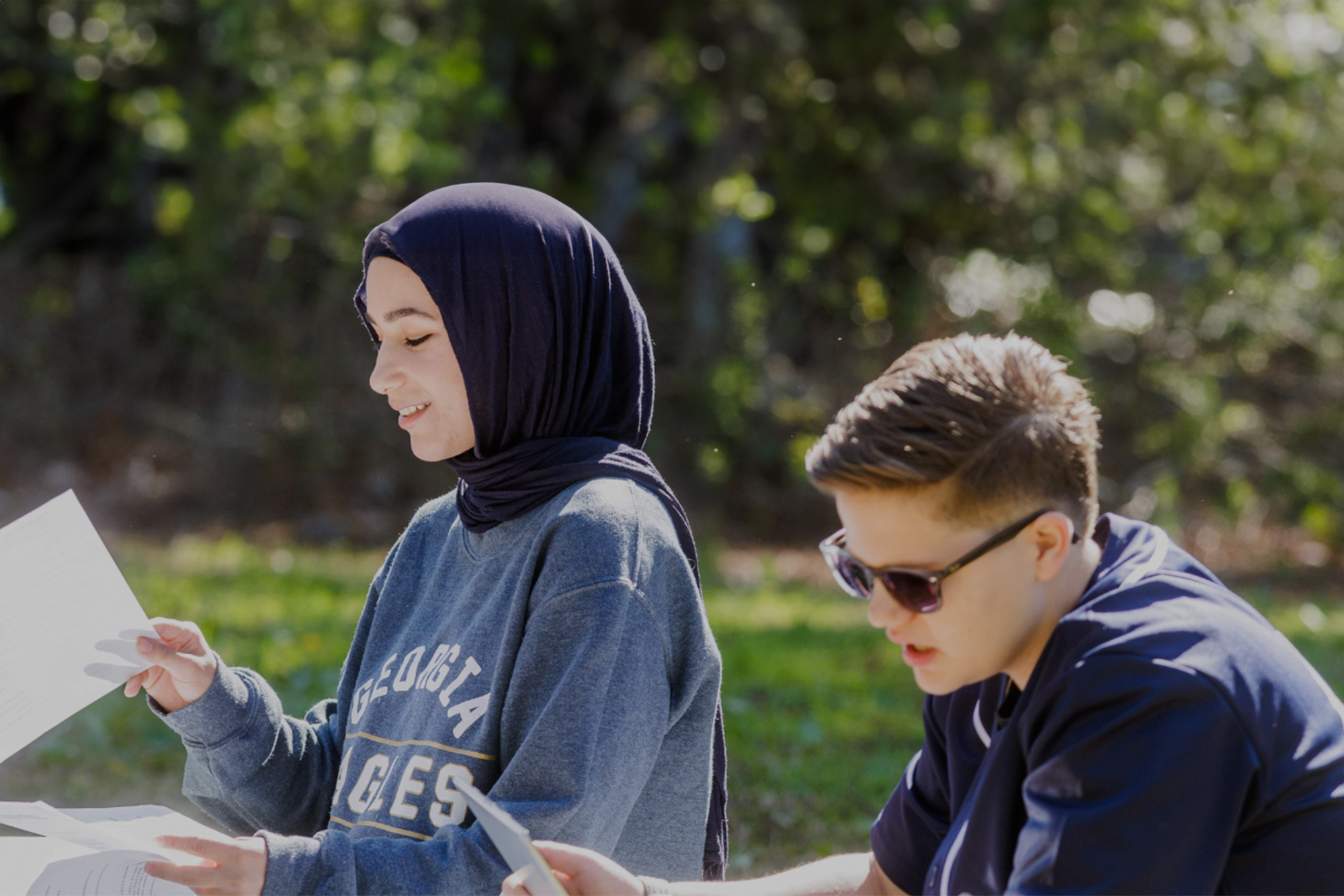 Two students sitting outside, studying together. One wears a headscarf and sweatshirt, holding papers; the other wears sunglasses and a dark shirt, holding a notebook.