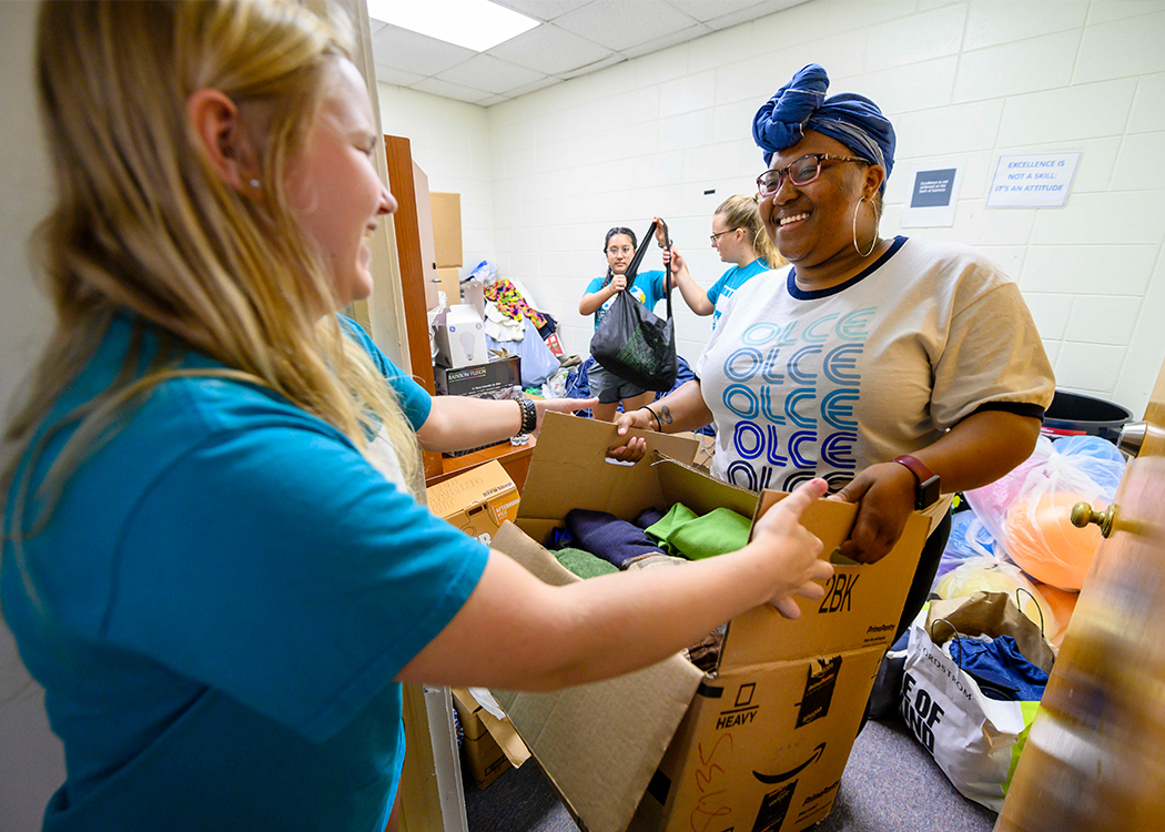 Two women smile as they hold a box of clothes together in a room filled with donated items. Another person in the background waves and smiles, suggesting a friendly, collaborative atmosphere.
