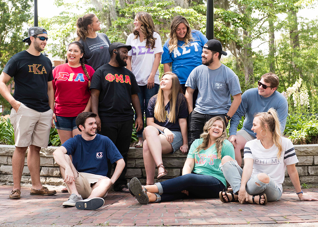 A diverse group of young adults wearing Greek letter shirts sit and stand together outdoors, smiling and talking in front of a brick path and greenery.