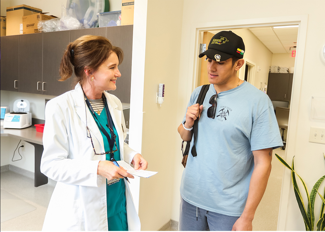 A female doctor in a white coat smiles and talks to a young man in casual clothes and a cap, who is carrying a backpack, inside a medical office or clinic.