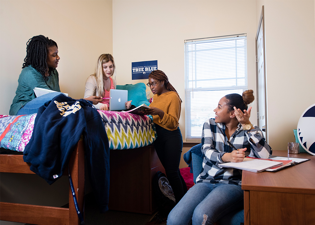 Four college students study together in a dorm room. Two stand by a lofted bed with laptops and papers, one sits on the bed, and another sits at a desk with a notebook, engaging in conversation.