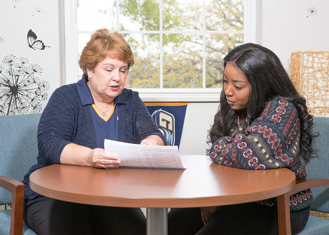 Two women sit at a round table indoors, one with short hair showing a document to the other with long hair. They appear to be discussing paperwork in a bright room with a window and wall decorations.