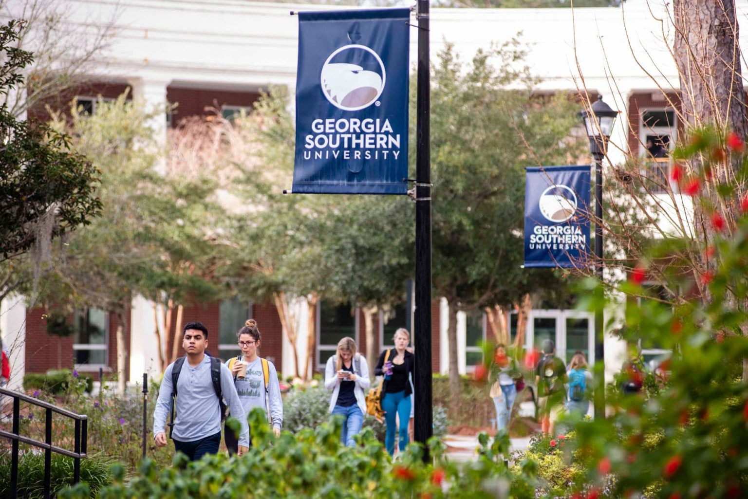 Students walk along a campus pathway lined with trees and bushes, passing by banners that read “Georgia Southern University.”