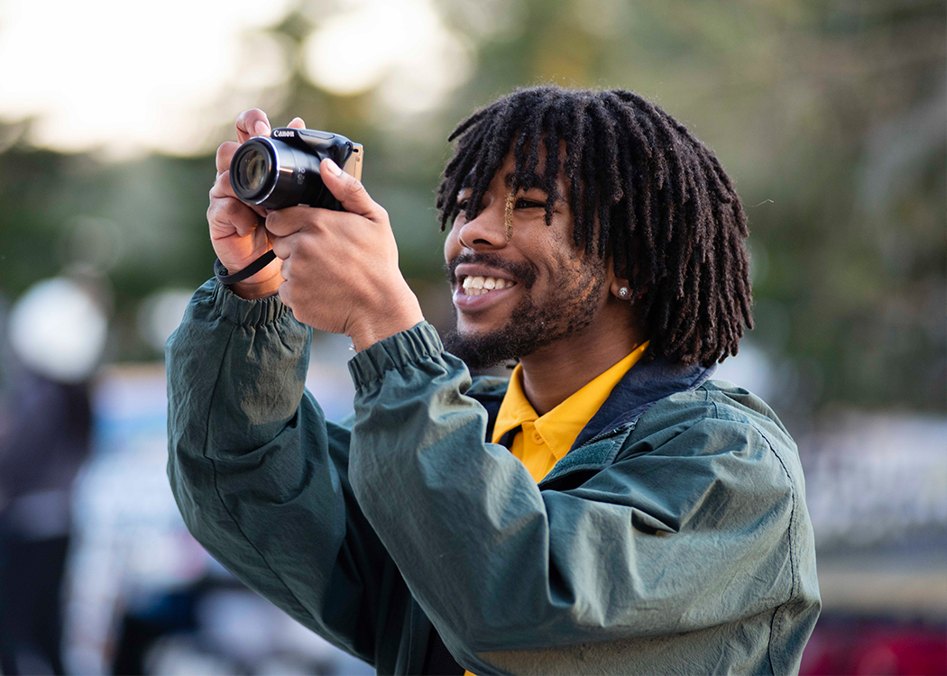 A young man with dreadlocks, wearing a green jacket and yellow shirt, smiles as he holds up a camera to take a photo outdoors.