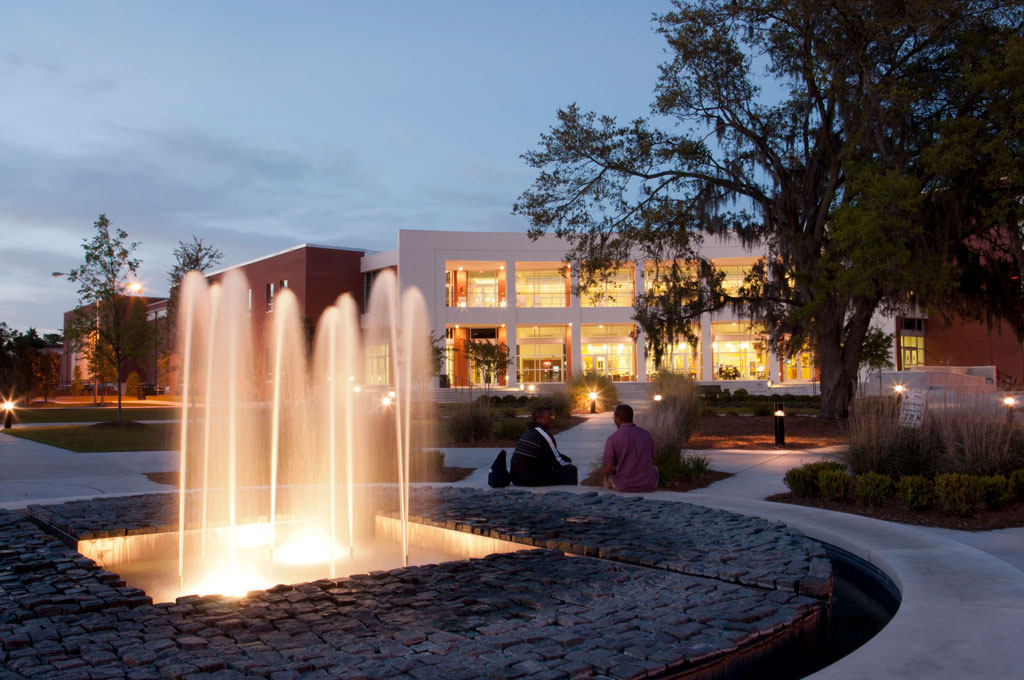 Two people sit near a lit fountain at dusk on a campus, with a modern building featuring large windows glowing warmly in the background, surrounded by trees and landscaped paths.