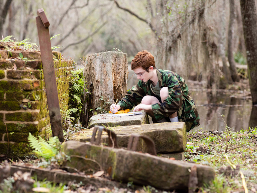 A young person in a camouflage jacket kneels outdoors, drawing or writing on a stone near old brick ruins and a wooded, swampy area. The scene is peaceful and natural with moss and ferns around.