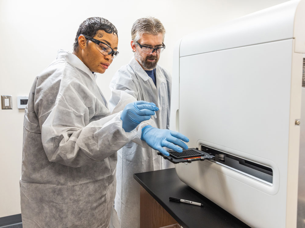 Two scientists wearing lab coats, gloves, and safety glasses place a tray with samples into a large laboratory machine in a bright, modern lab setting.