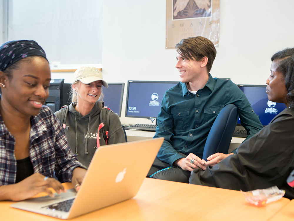 Four students sit together in a computer lab, talking and smiling. One student types on a laptop while the others engage in conversation. Computer monitors and a poster are visible in the background.