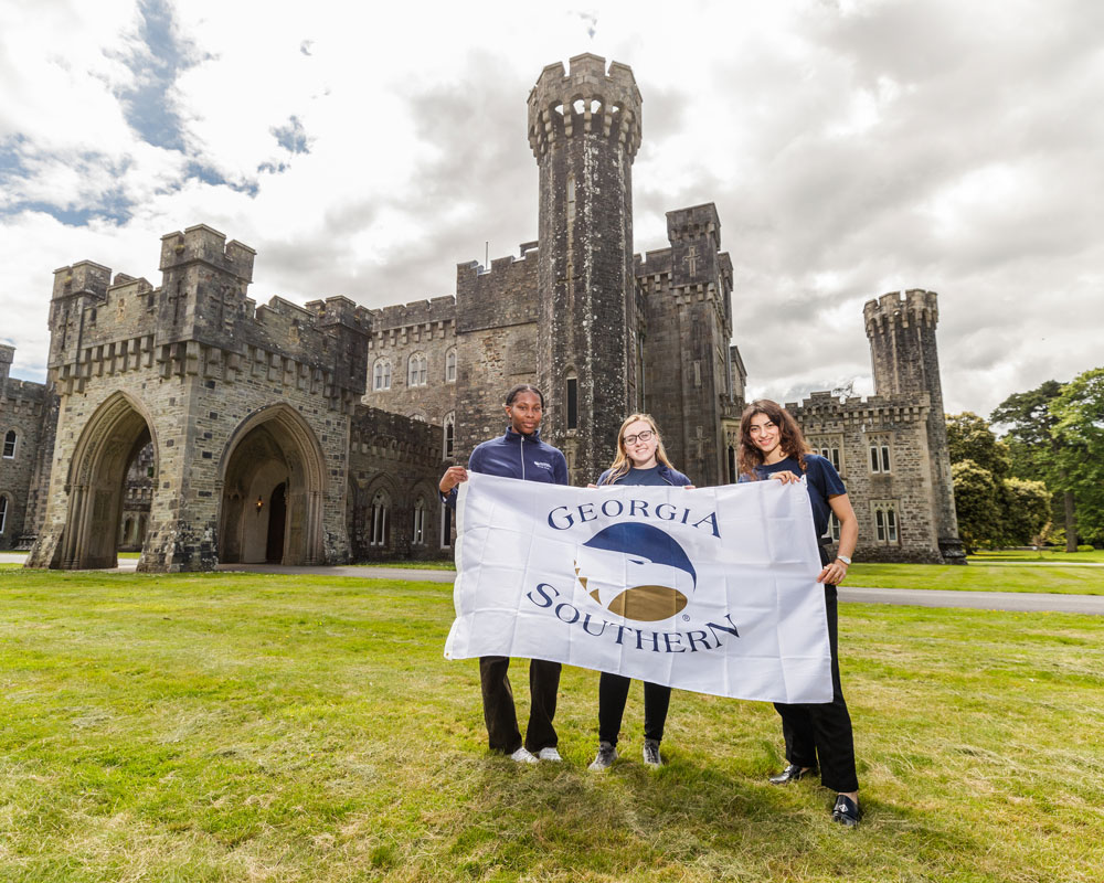 Three people stand on grass in front of a stone castle, holding a white Georgia Southern flag with a blue eagle logo.