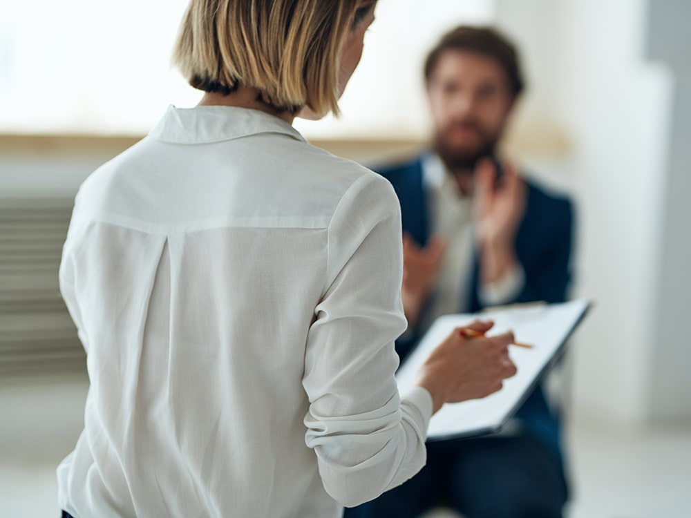 A woman takes notes as an out of focus man speaks to her.