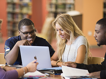 Four students sit together at a table in a library, studying and collaborating while looking at a laptop and books.