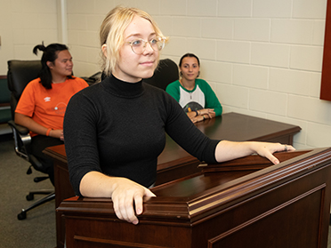 A smiling young woman stands at a wooden podium in a classroom, while three people sit at desks behind her, listening attentively.