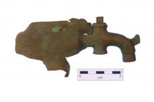 An irregularly shaped brass object approximately 12 centimeters long. One end is shaped like a faucet tap. The other end is flat with curled ragged edges like it was torn from a larger object.