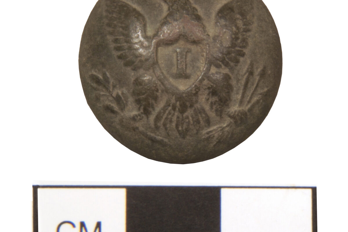 A metal button embossed with an eagle carrying an olive branch and arrows in its talons. The button is about 70 millimeters in diameter.