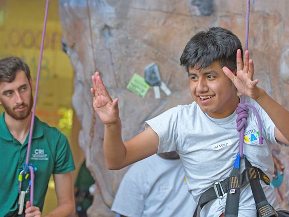A researcher and camp leader watches closely as a boy swings hands-free in a rock-climbing harness.