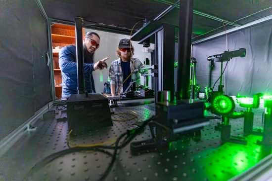 An instructor explains something to a student as they observe the operation of a large piece of equipment that glows bright with green laser light. Both are wearing safety shades.