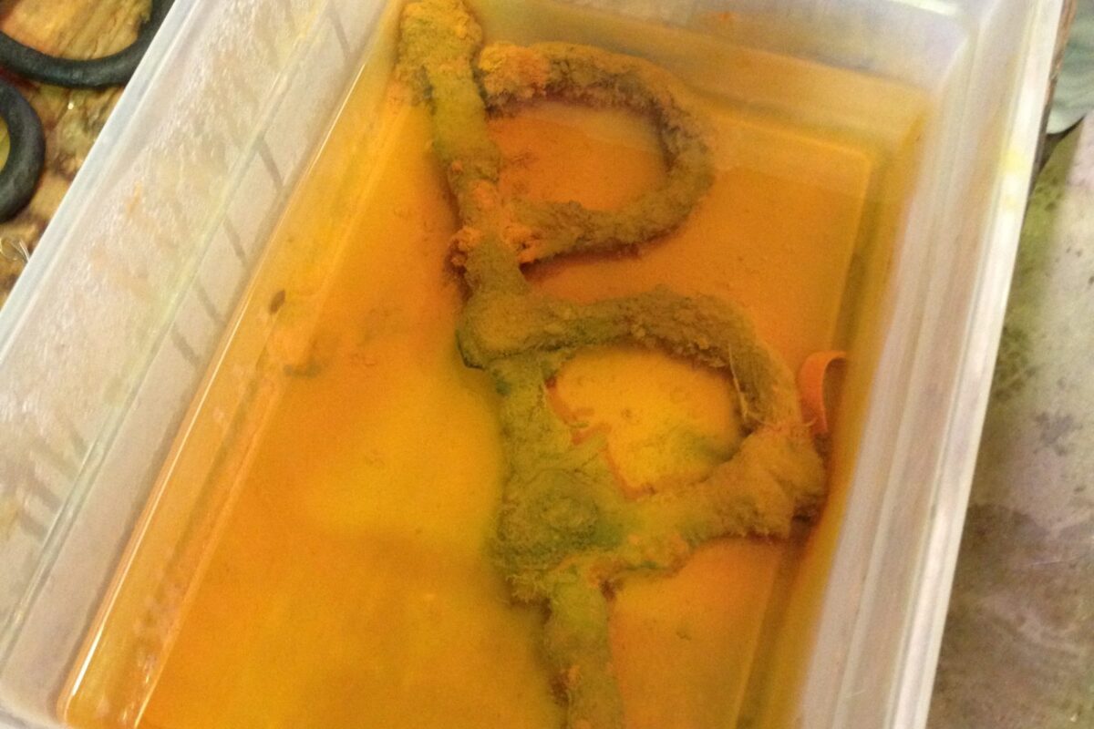 A pair of corroded shackles in a plastic tub in yellow liquid.
