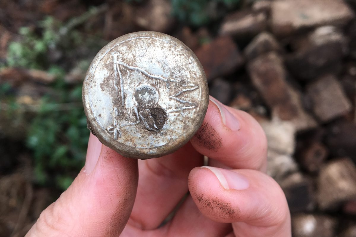 A person holds a small object with a circular metal top. The metal is embossed with a flag logo.