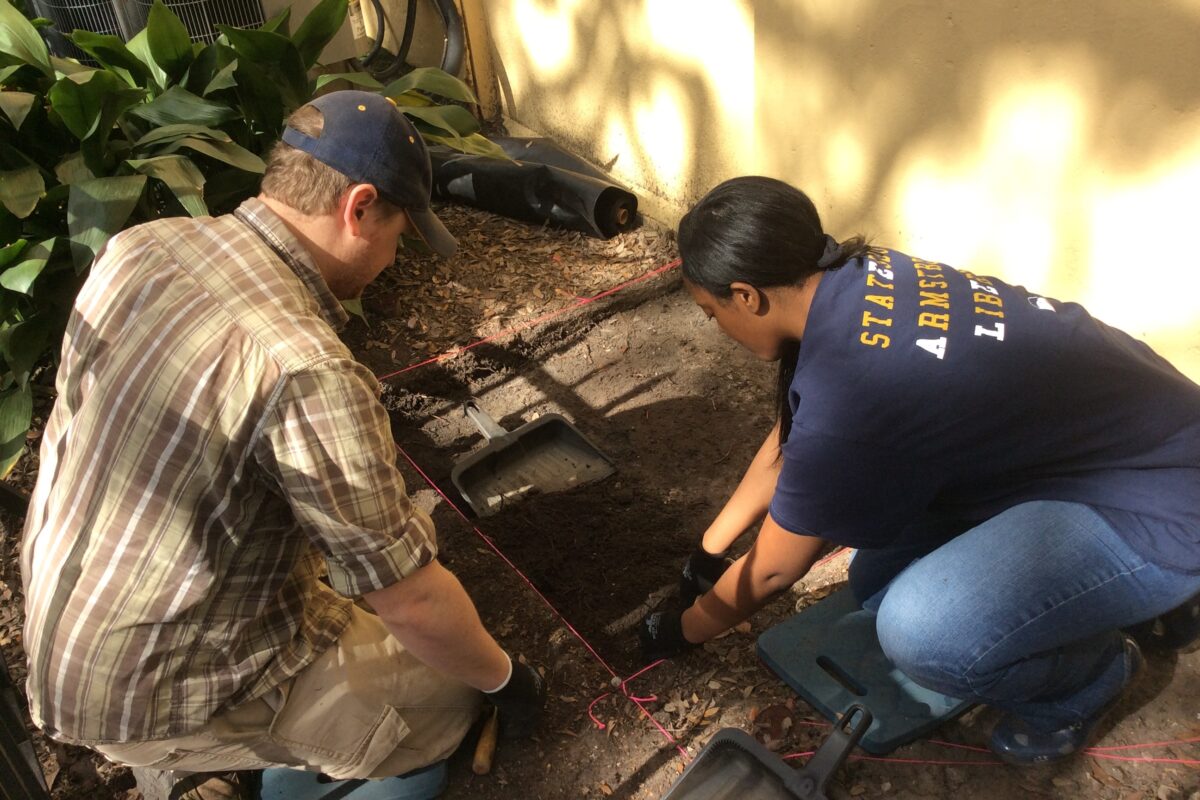 A man and woman collect a sample of topsoil from a marked-off square near the utility entrance of a building.