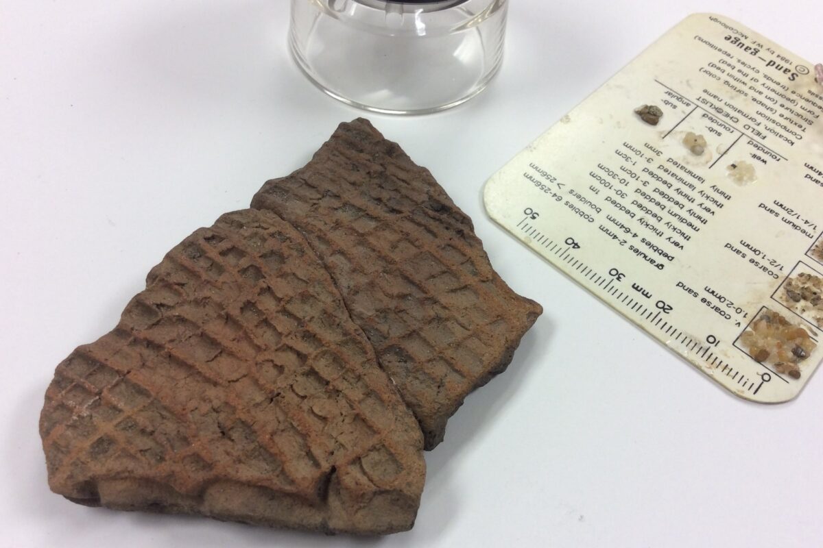 A small fragment of rough pottery, roughly 4cm by 7cm, with a pattern of square blocks stamped in it. The photo also includes a 10x magnifying loupe and an information card with a 50mm ruler.
