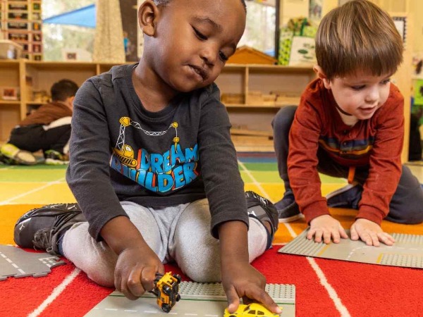 Two young children playing with toy cars on a brightly colored carpet in a childcare facility.
