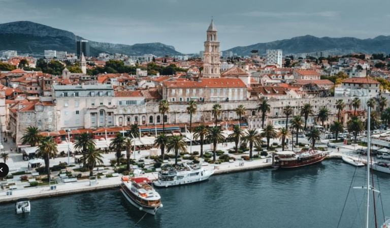 A view of a city from the harbor with red roofed white buildings and a few tall steeples. There are mountains in the background and palm trees along the shoreline.